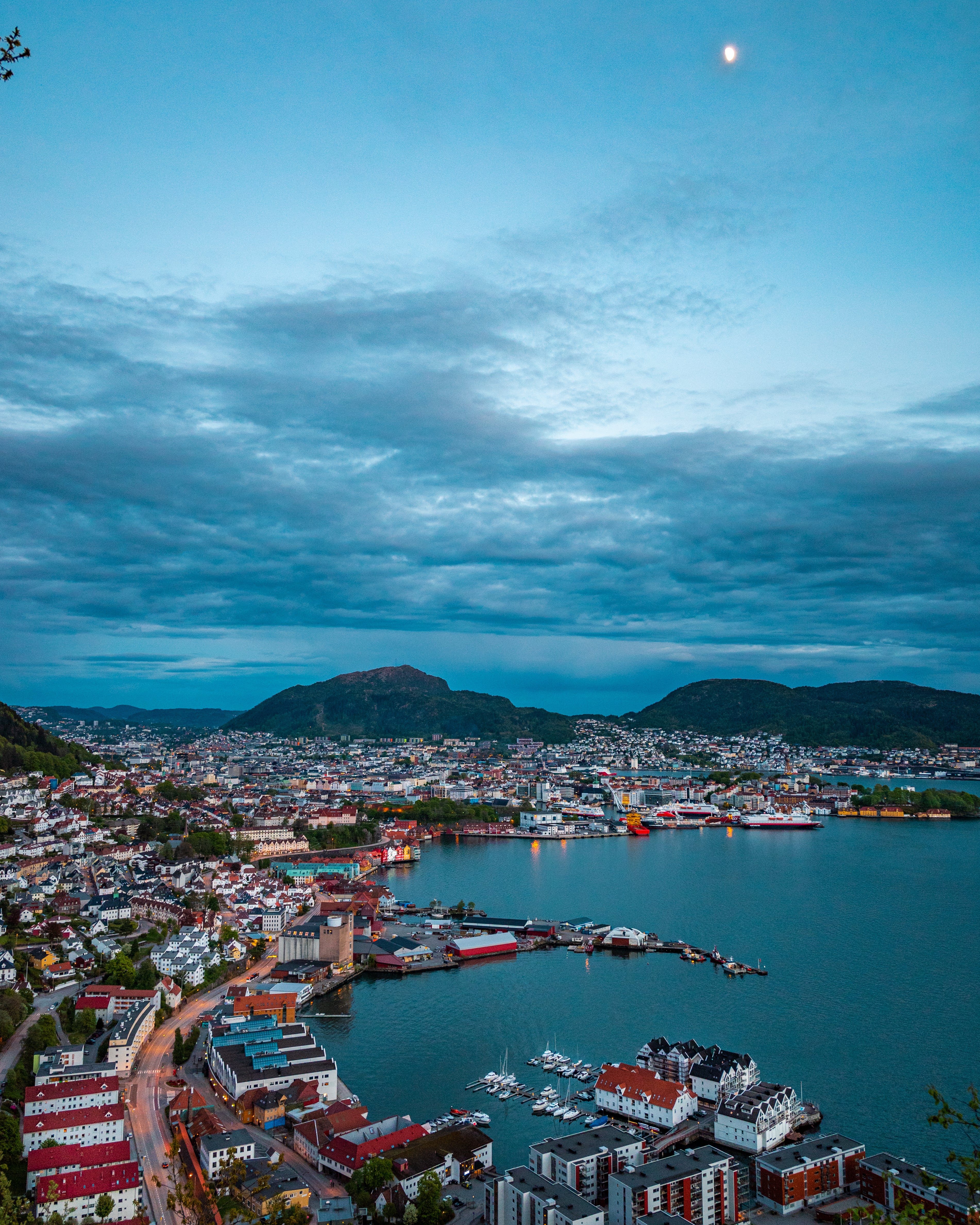 A wide panoramic view of Bergen, Norway, at dusk. The city lights twinkle along the harbor and hillside, with dark mountains and a cloudy blue sky in the background