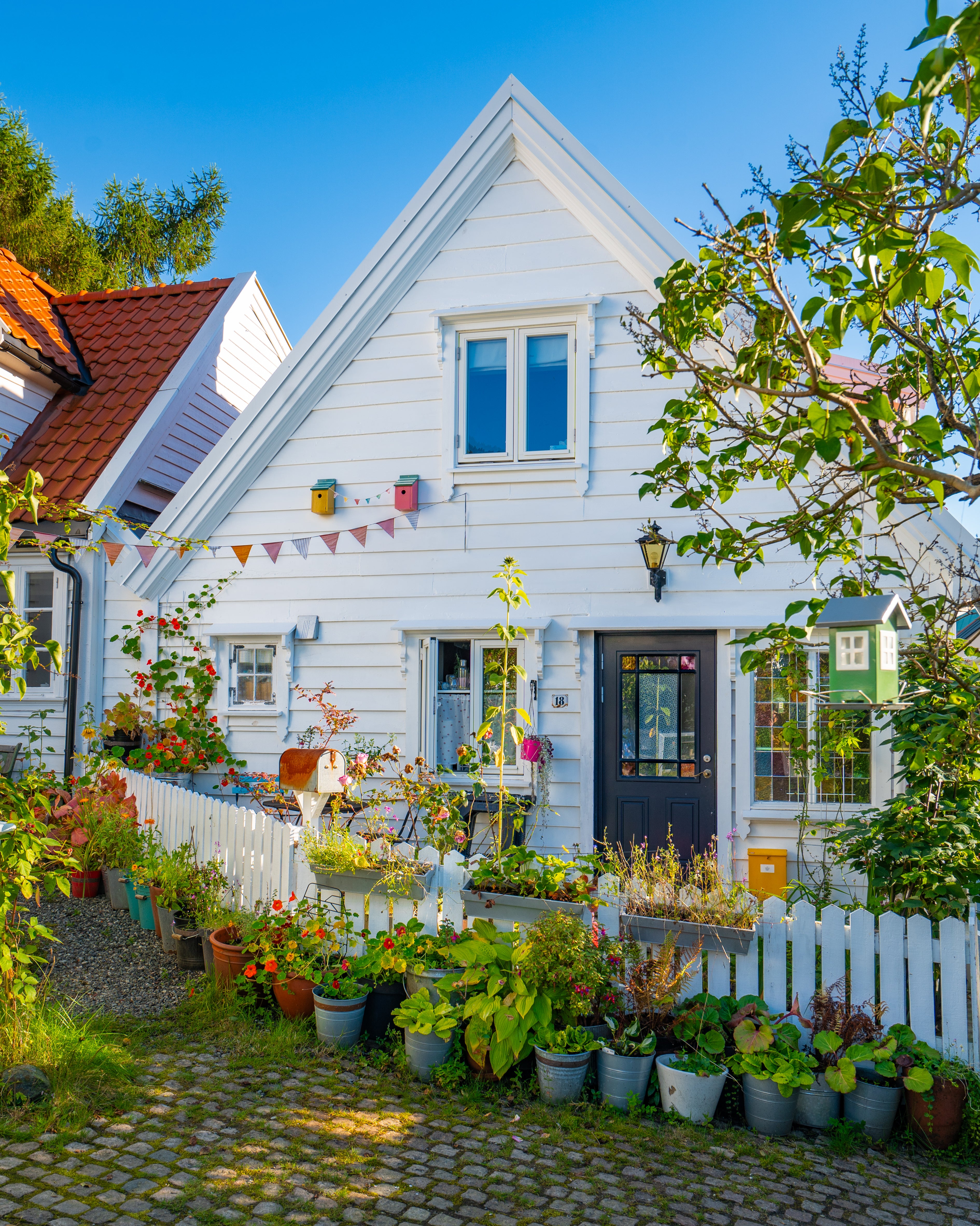 A charming, white-painted traditional wooden house in Bergen, Norway, decorated with colorful flowers, birdhouses, and bunting under a clear blue sky.