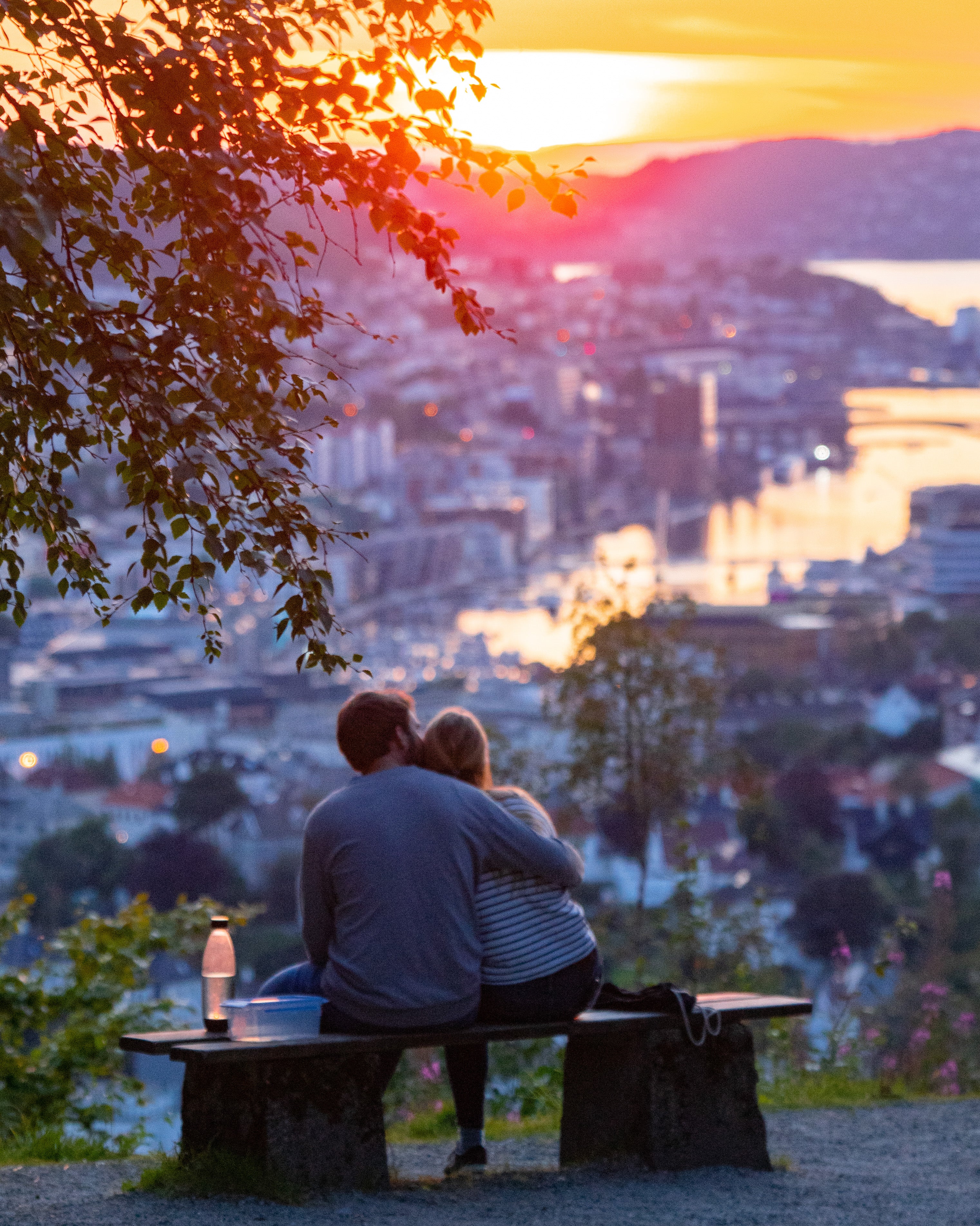 A couple sitting on a wooden bench at a viewpoint, embracing while watching a vibrant pink and orange sunset over the city and harbor of Bergen, Norway.