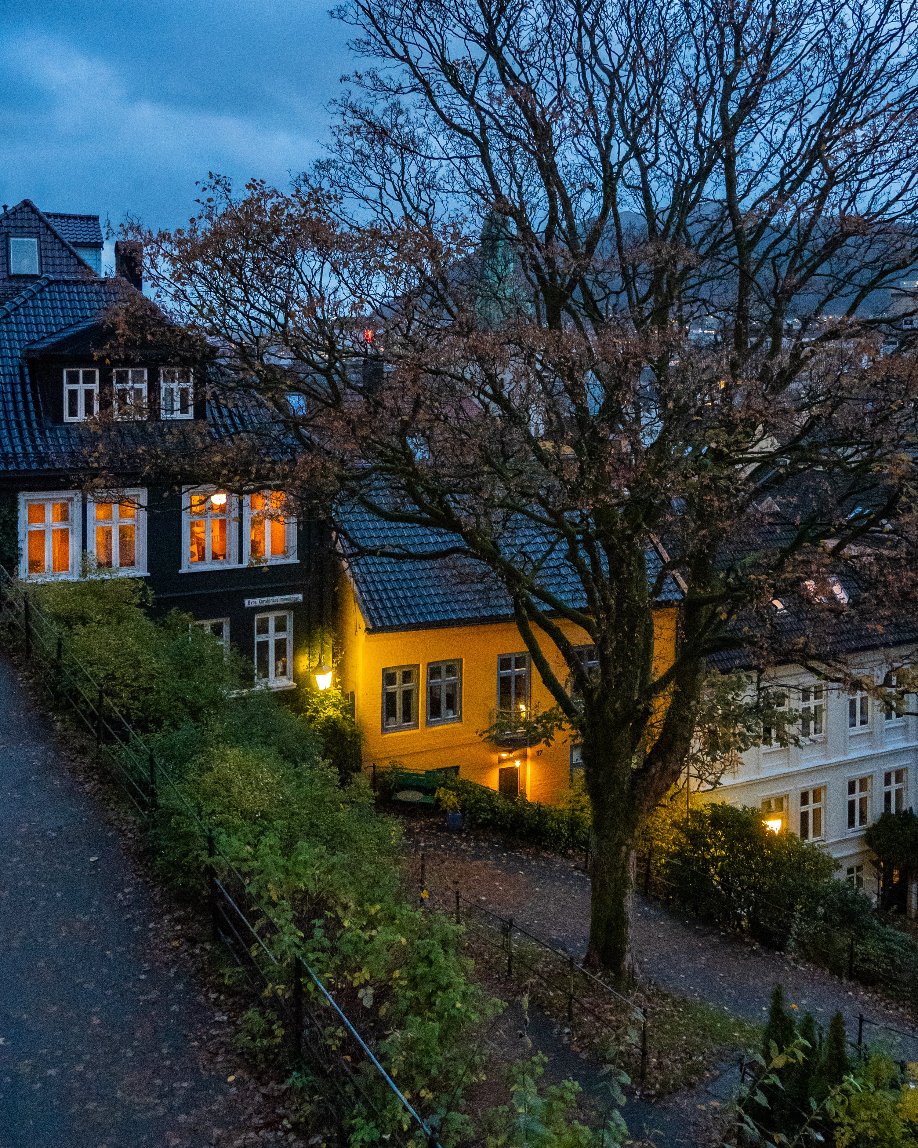A cozy evening scene in a Bergen hillside neighborhood at dusk. Warm yellow lights shine from the windows of traditional black, yellow, and white wooden houses, surrounded by autumn trees and a quiet pedestrian path