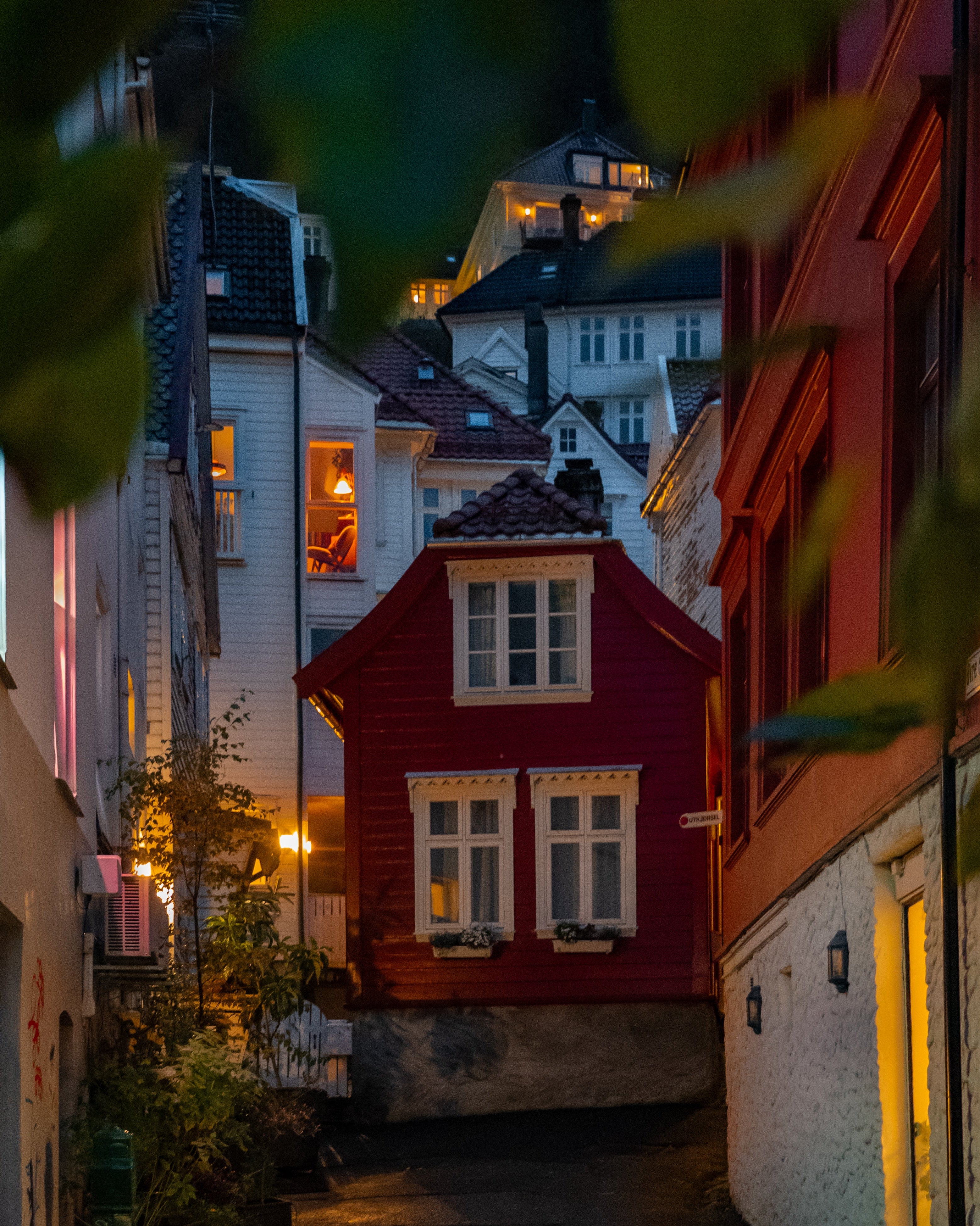 A narrow, atmospheric street in Bergen, Norway, at night. A small red traditional wooden house stands at the end of the alley, with warm lights glowing from windows in the surrounding white buildings