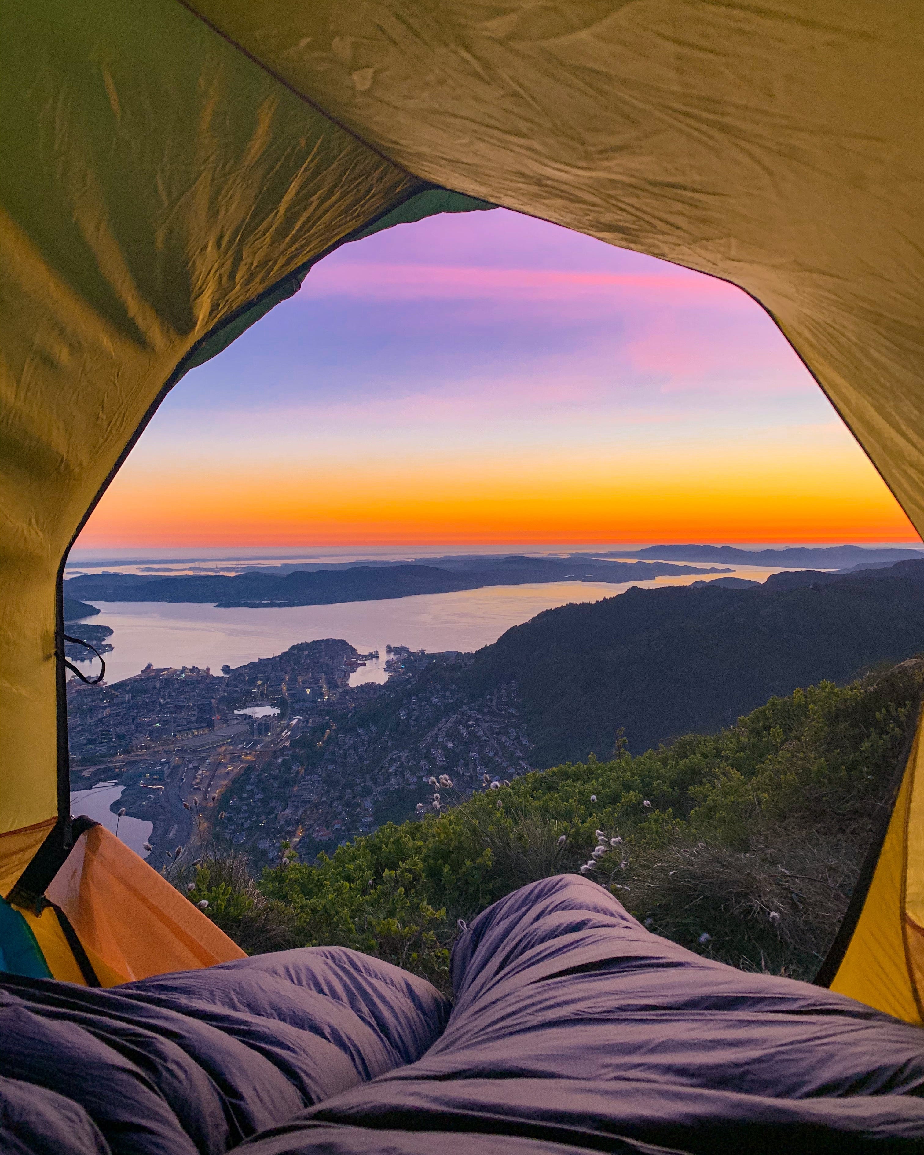 A breathtaking sunset view of Bergen city and the Norwegian fjords seen from the inside of a tent on Mount Ulriken. The sky is vibrant purple and orange, framing the coastal landscape.