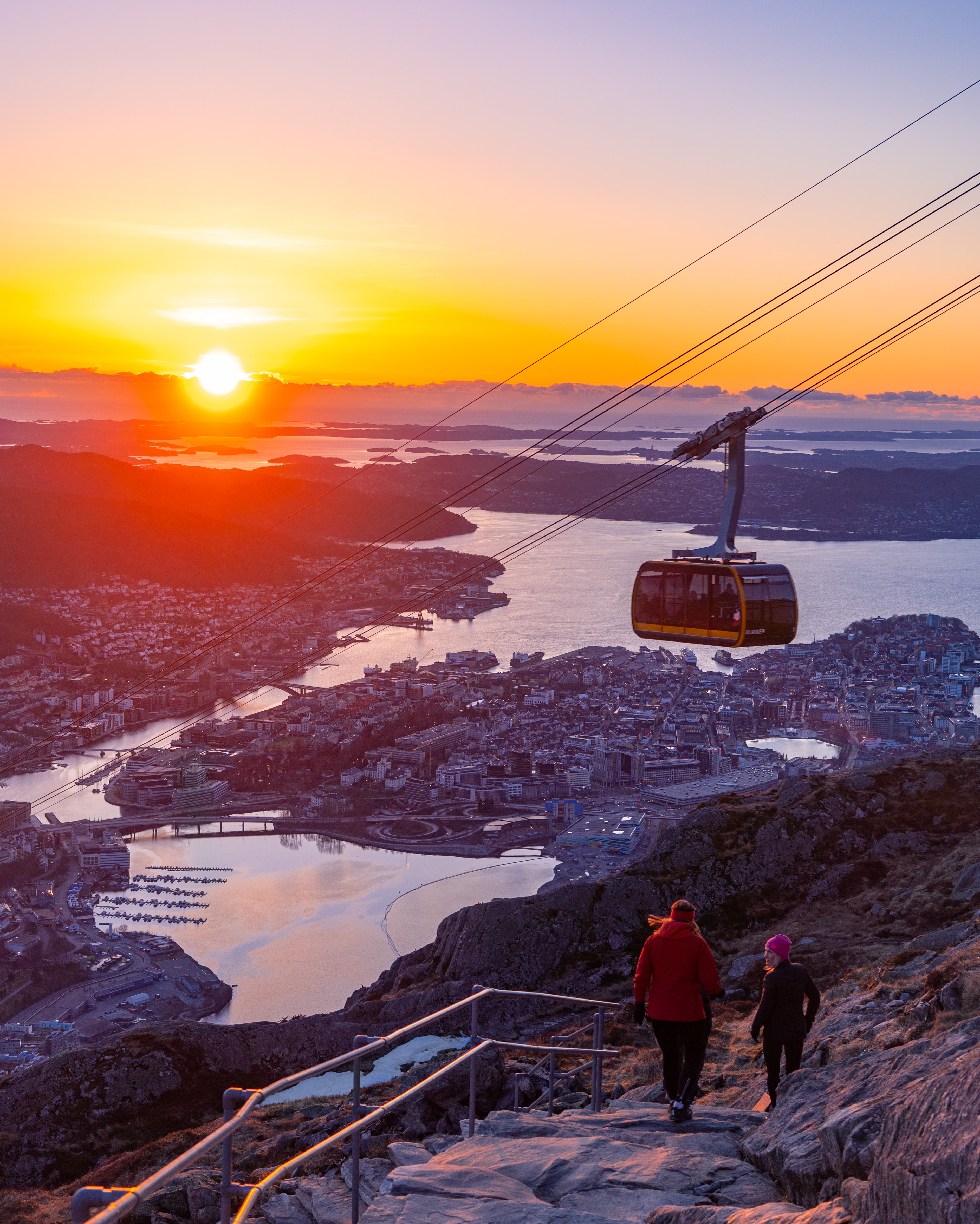 The Ulriken cable car (Ulriksbanen) suspended over Bergen city, Norway, during a golden sunset. Two hikers are walking down a stone path in the foreground, with the city and fjords stretching out in the background.