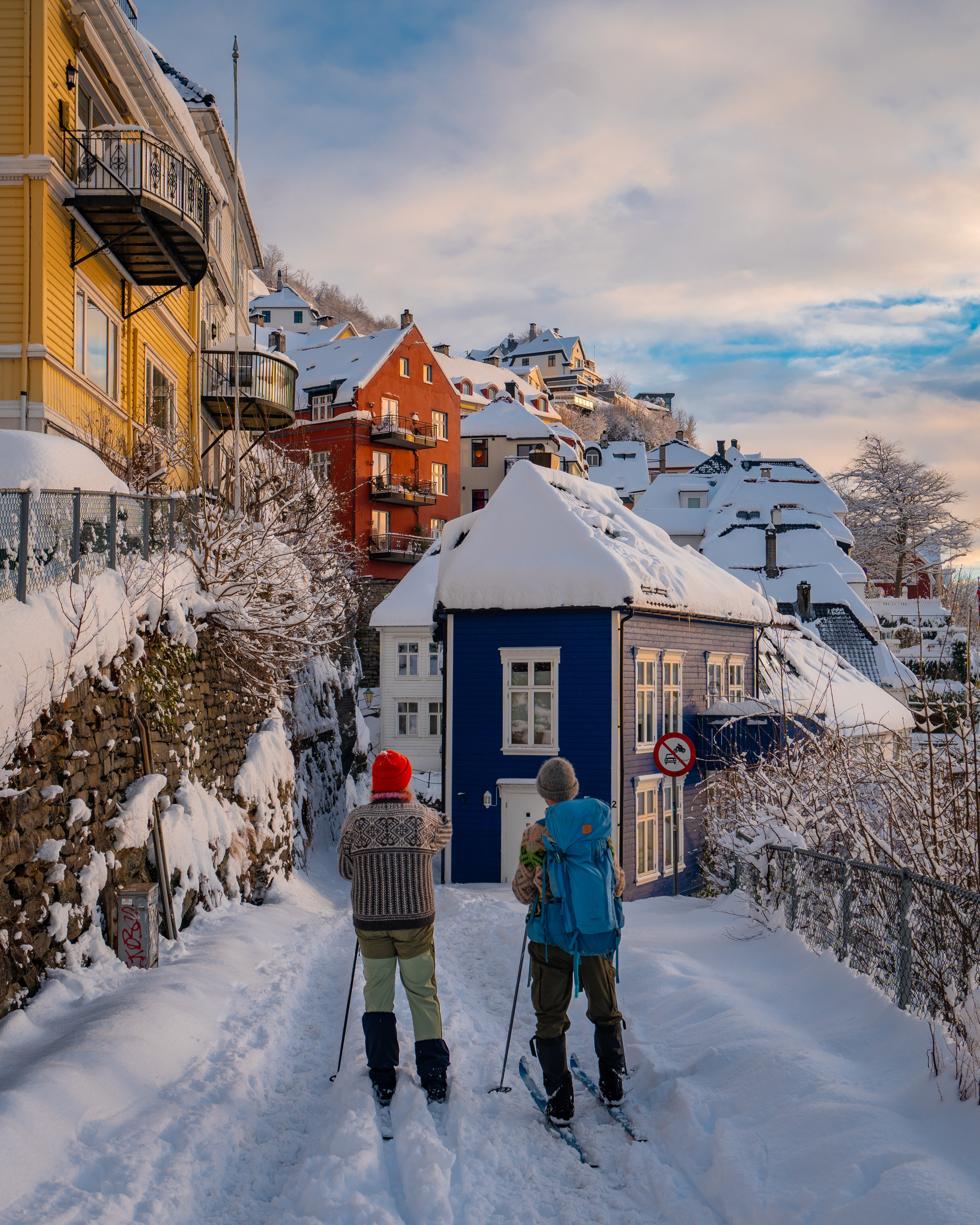 Two people on cross-country skis exploring the snow-covered, narrow streets of Bergen, Norway, surrounded by colorful traditional wooden houses under a winter sky.