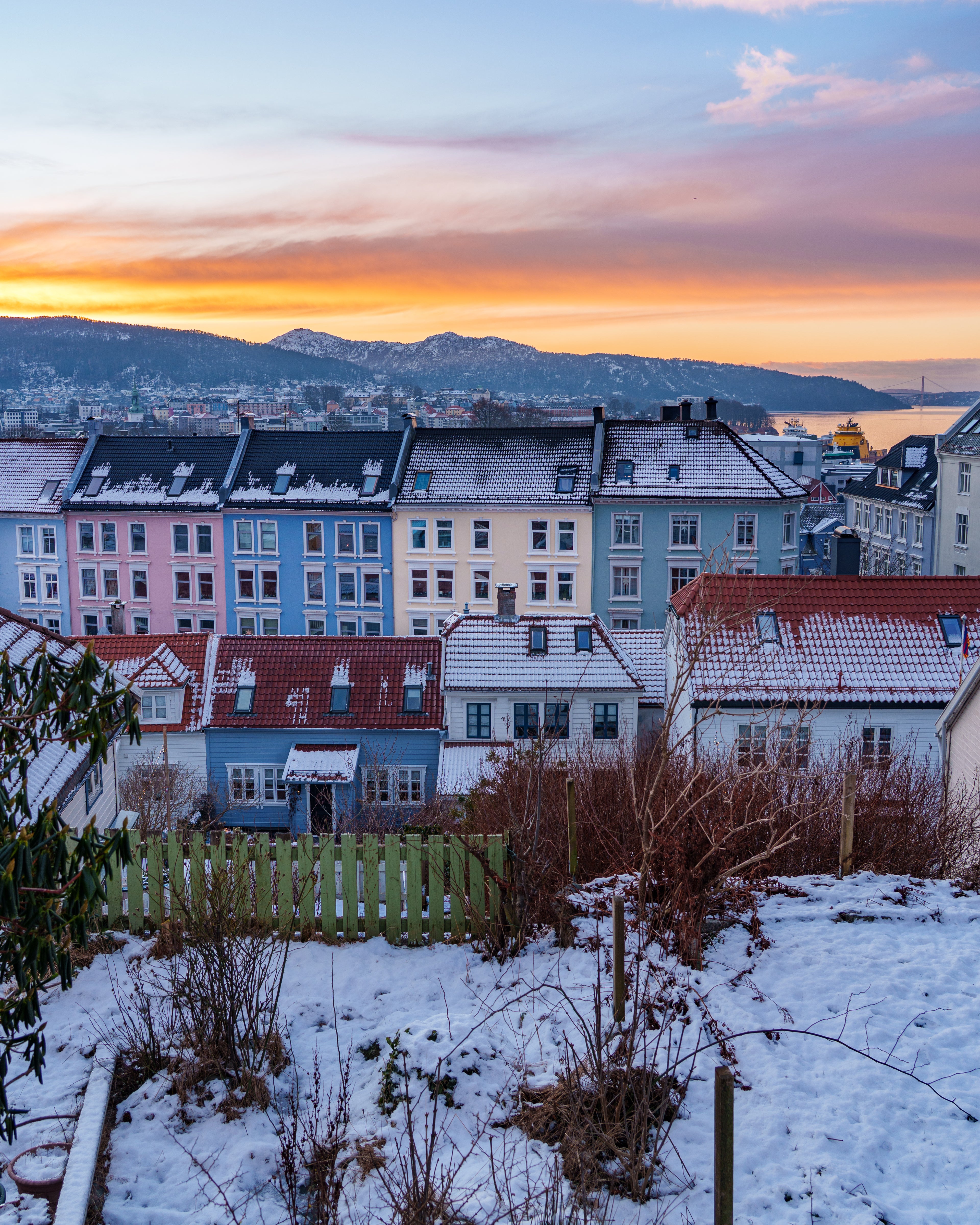 Rows of colorful historic houses in pink, blue, and yellow covered in snow under a vibrant orange and purple sunset sky in Bergen, Norway, with snow-capped mountains in the distance